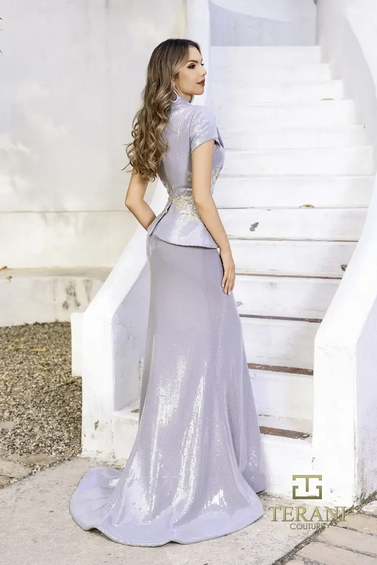 Woman in a sleek, shimmering silver gown with gold embroidery stands confidently by white curving stairs. The scene conveys elegance and sophistication.