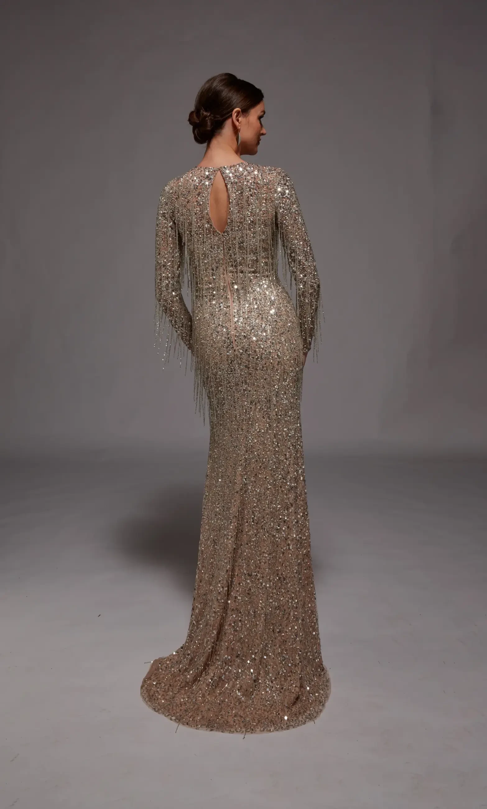 A woman poses gracefully in a shimmering, long-sleeved, gold sequin gown with fringe details and a high slit, exuding elegance and confidence.
