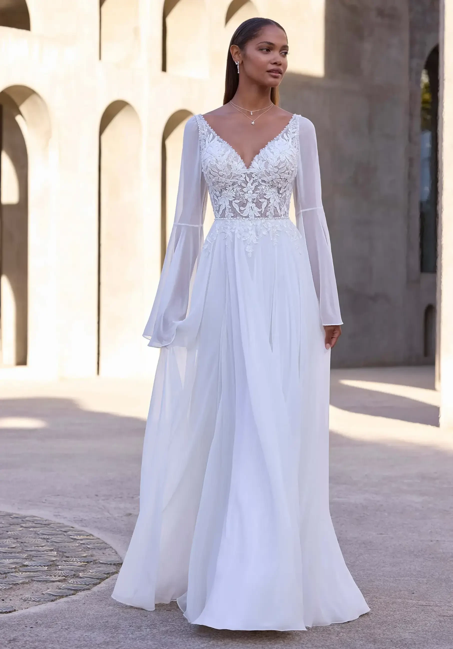Model is wearing a chiffon A-line wedding dress with a sequin and beaded V-neck bodice, low back, and detachable sheer bell sleeves.