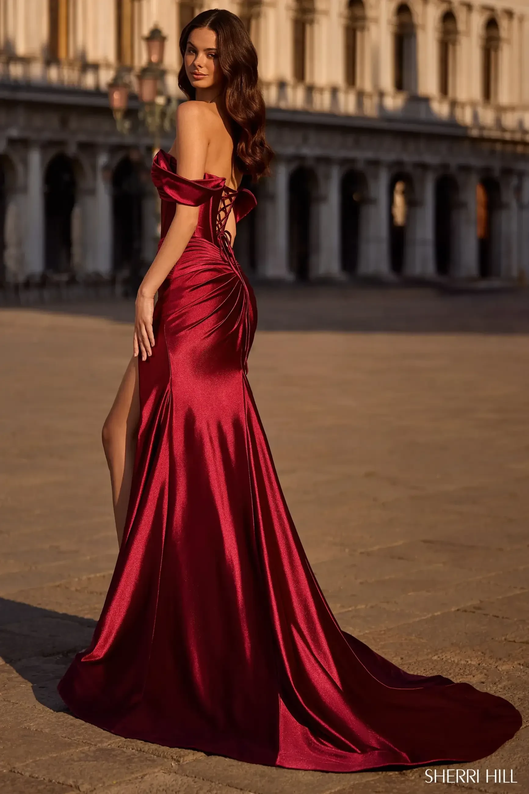 Model is wearing a satin Sherri Hill long fitted dress with a sweetheart neckline, off-the-shoulder cap sleeves, corset bodice, keyhole cutout, and side slit.