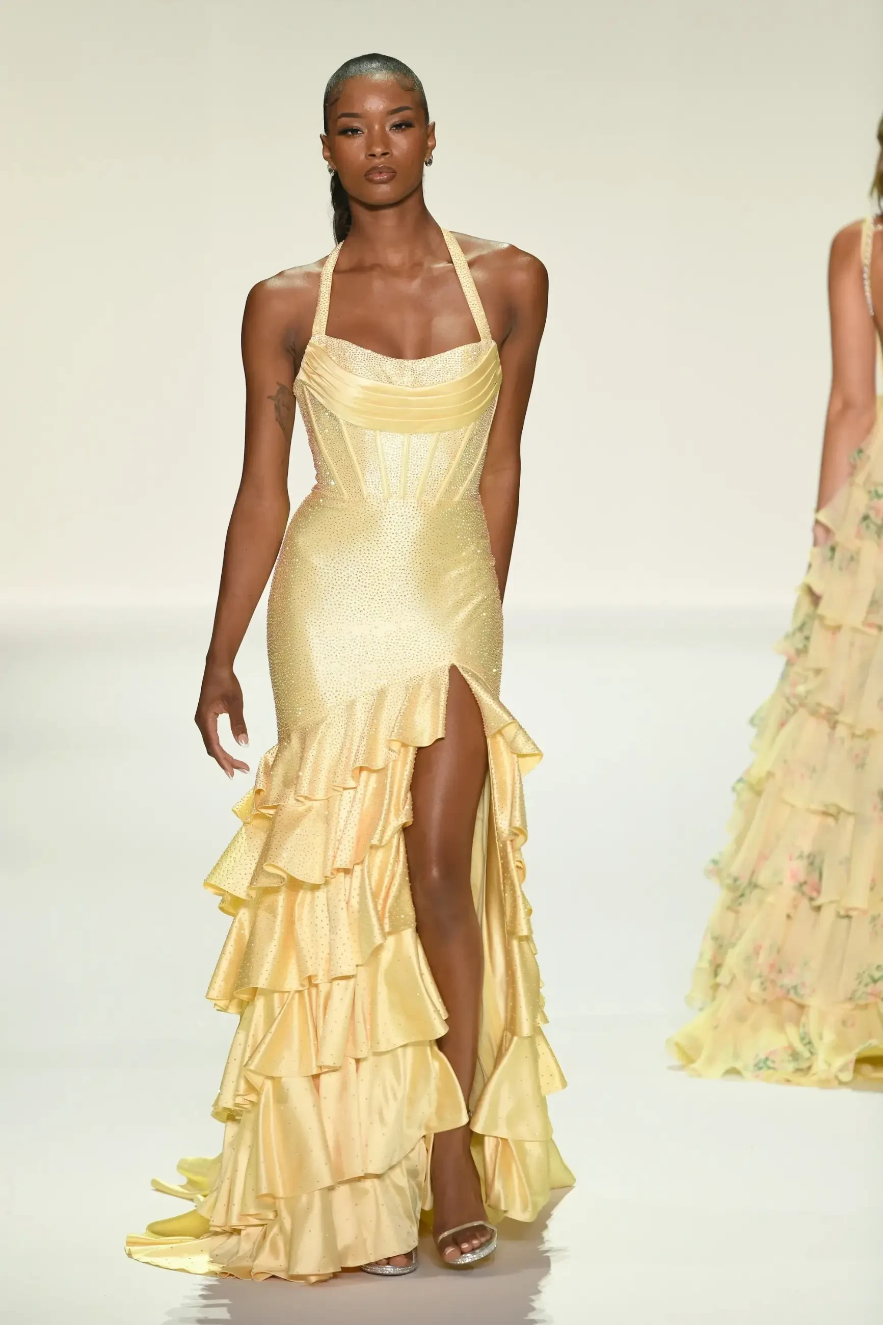 Model in a shimmering yellow halter-neck dress with ruffled layers and thigh-high slit walks confidently on the runway, exuding elegance and poise.