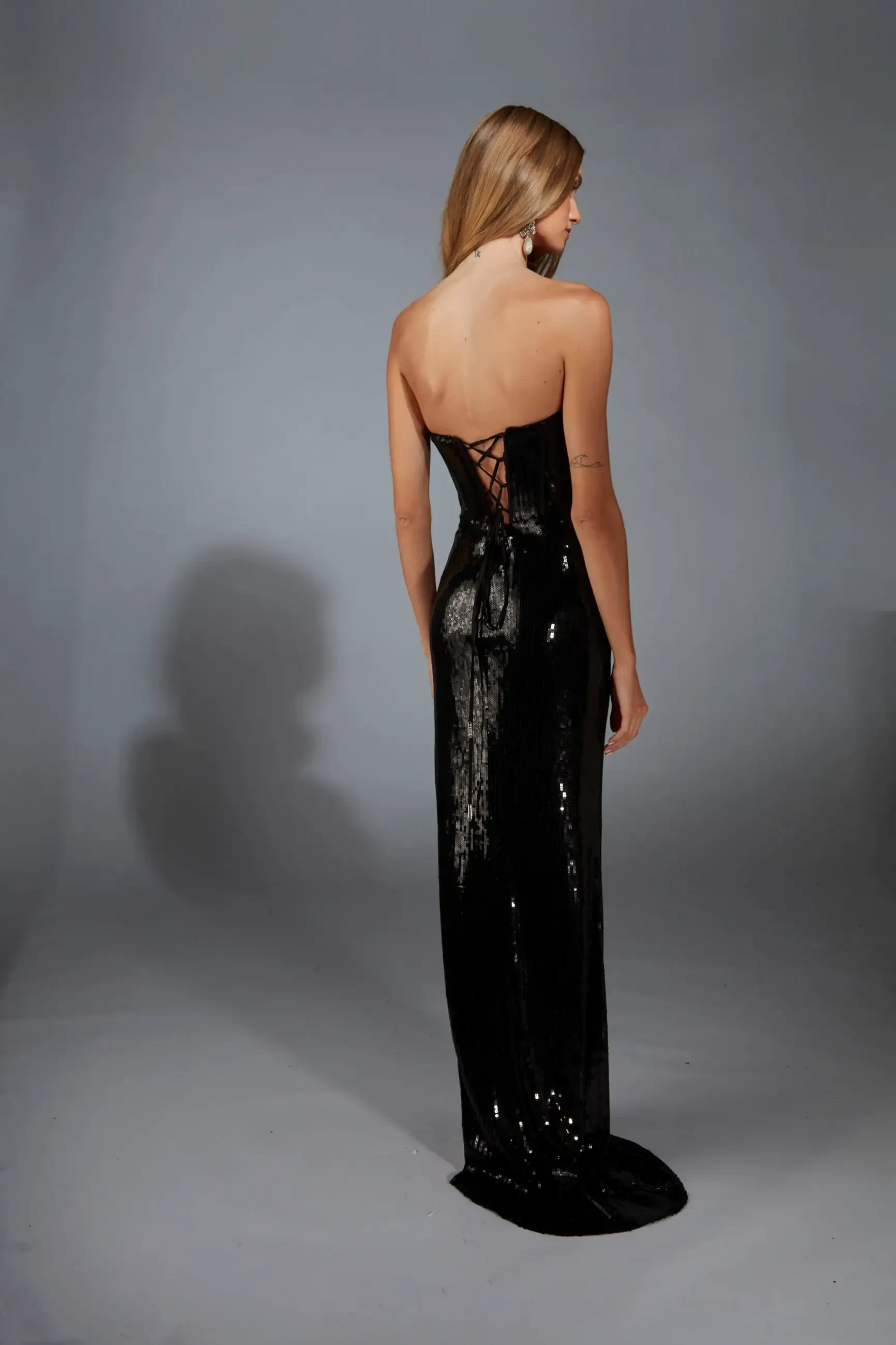 Model is wearing a fully sequined fitted gown with a strapless corset bodice, high slit, low lace-up back, and a sweep train.