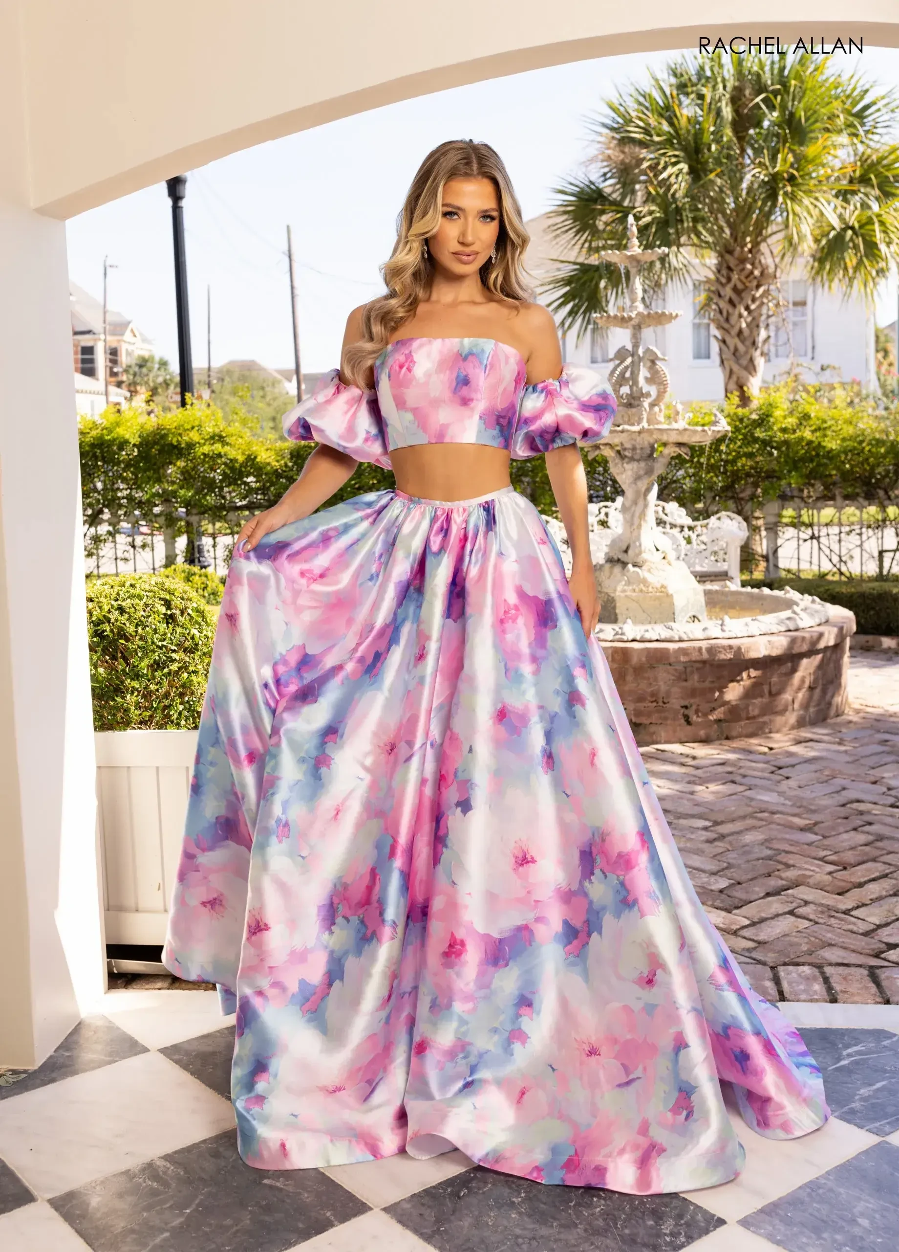 Woman in a vibrant floral two-piece gown with off-shoulder puff sleeves stands elegantly outdoors. She poses confidently near a fountain, exuding a summery, radiant vibe.