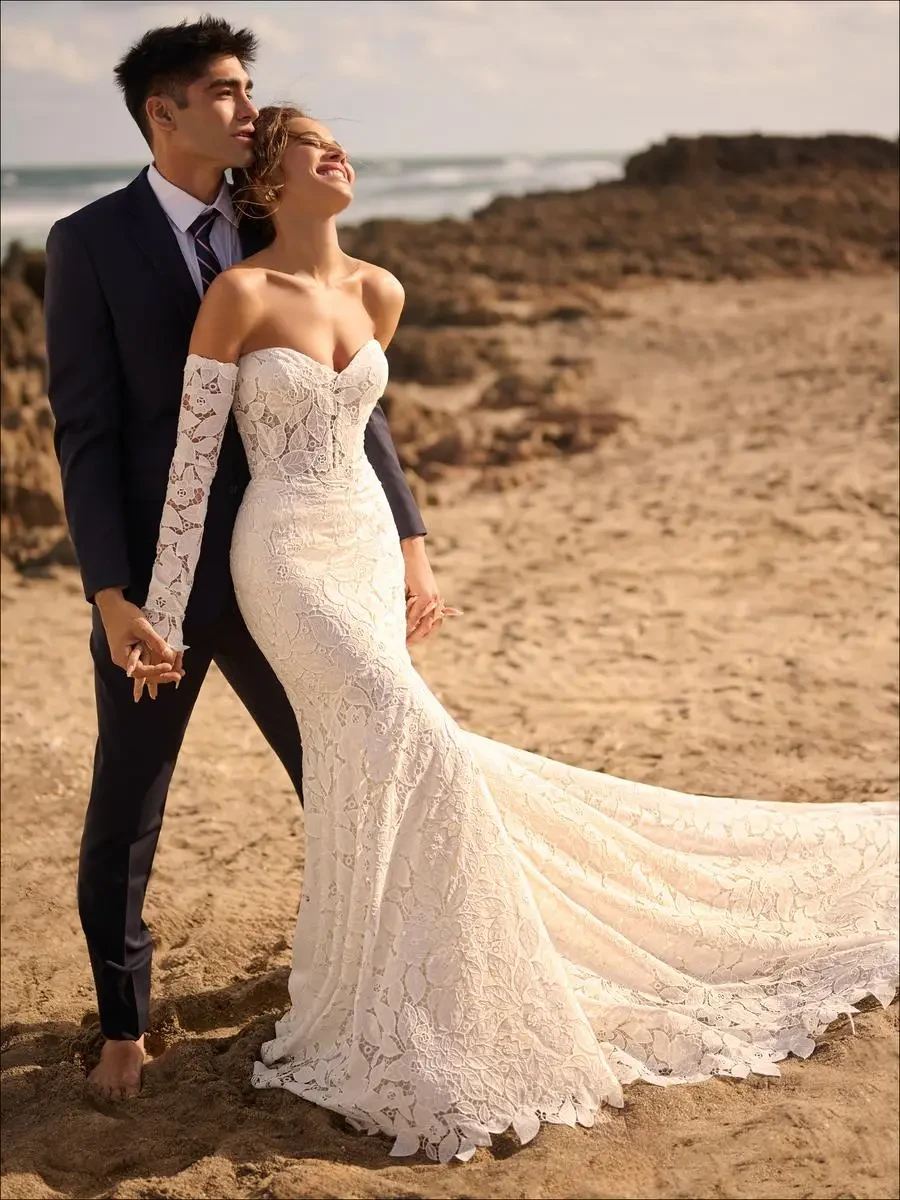 Bride and groom stand on a sandy beach. The bride wears a lace gown with a long train and looks joyfully upward. The groom holds her hand, gazing into the distance.