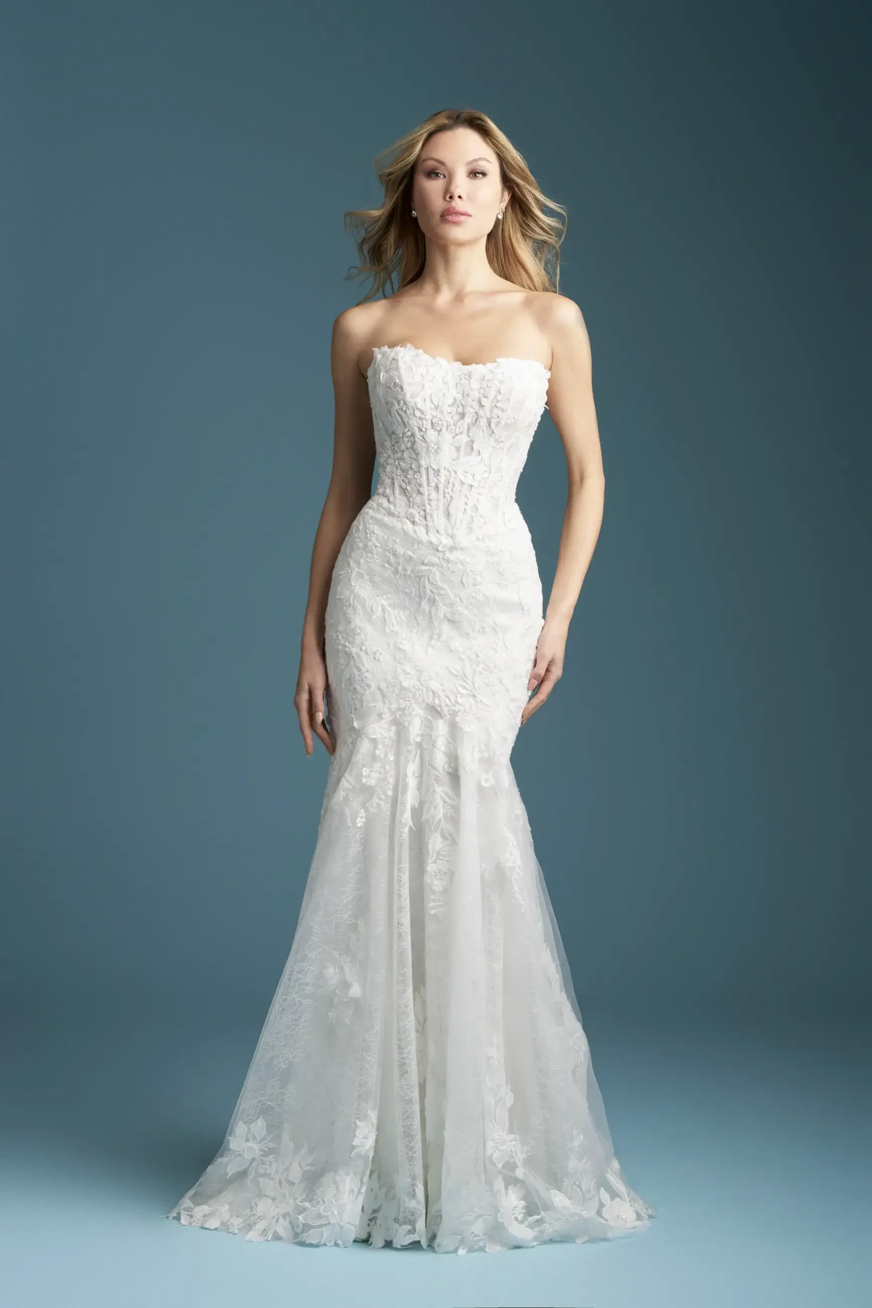A woman in a strapless, lace-embroidered white wedding gown stands gracefully against a muted blue background, exuding elegance and poise.