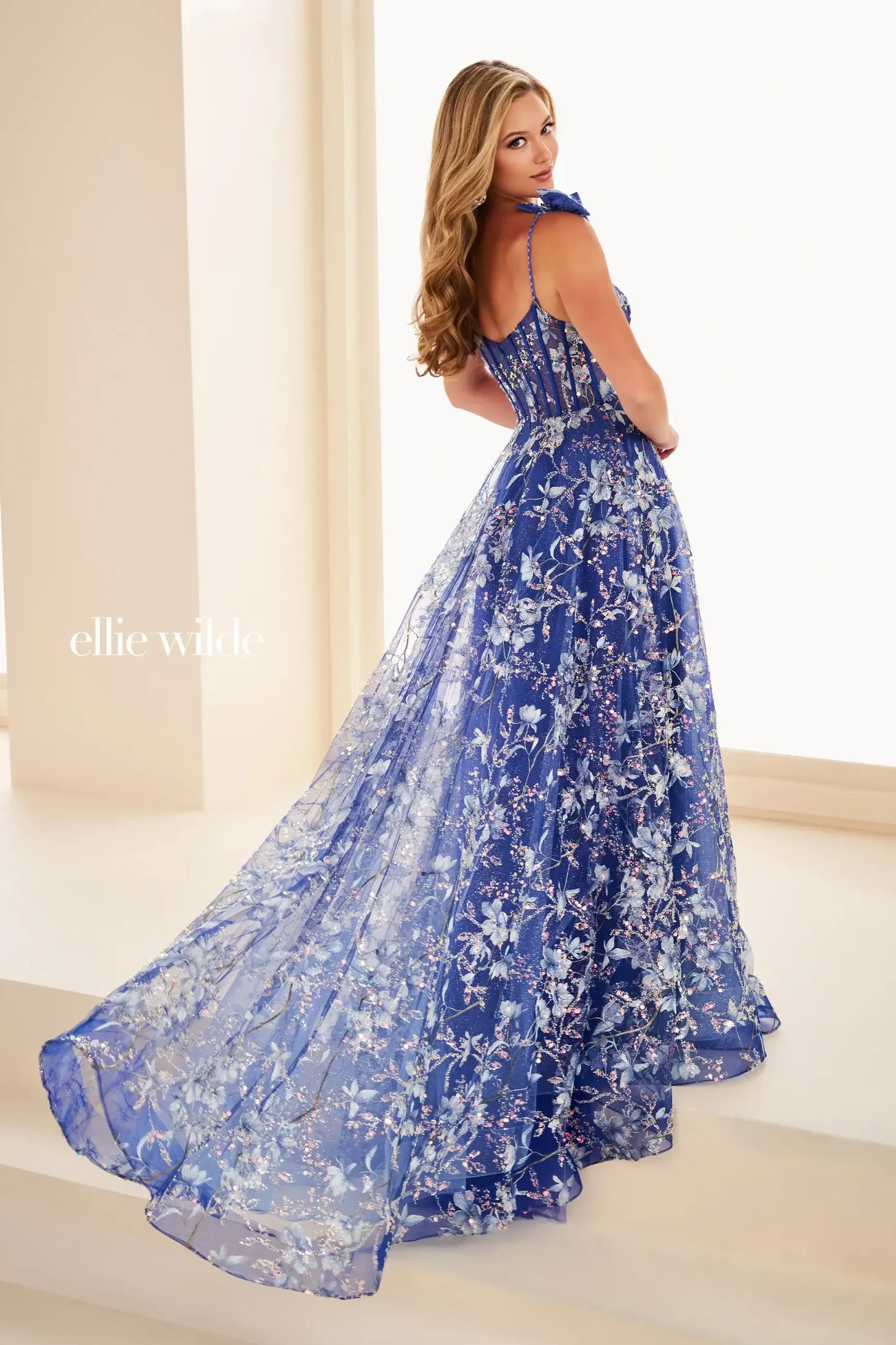 A woman in a flowing blue floral gown glances back, exuding elegance. The dress features intricate lace detailing, conveying a sense of grace and sophistication.
