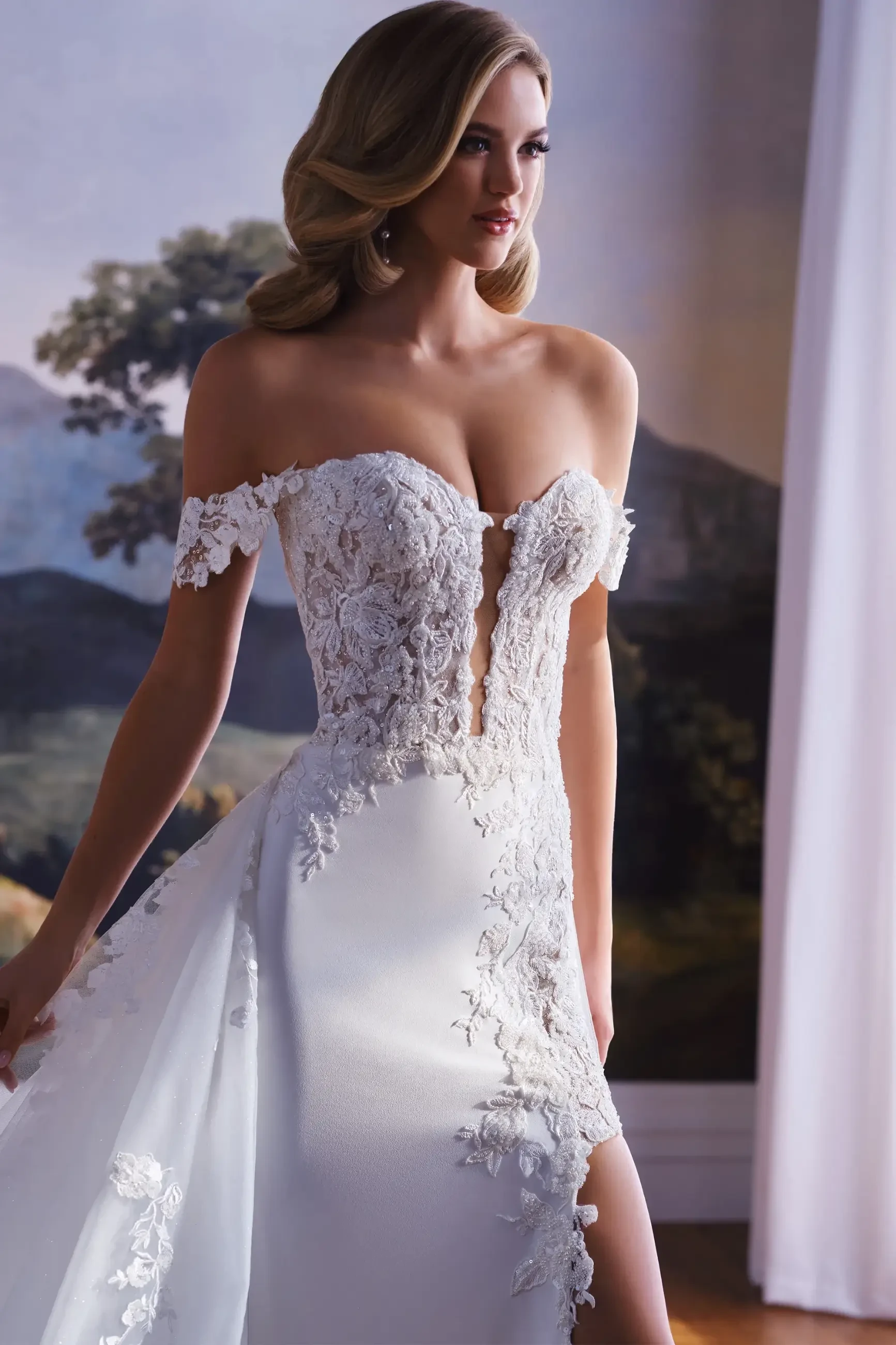 Model is wearing a beaded lace wedding gown with a plunging neckline, removable off-the-shoulder straps, and a detachable train.