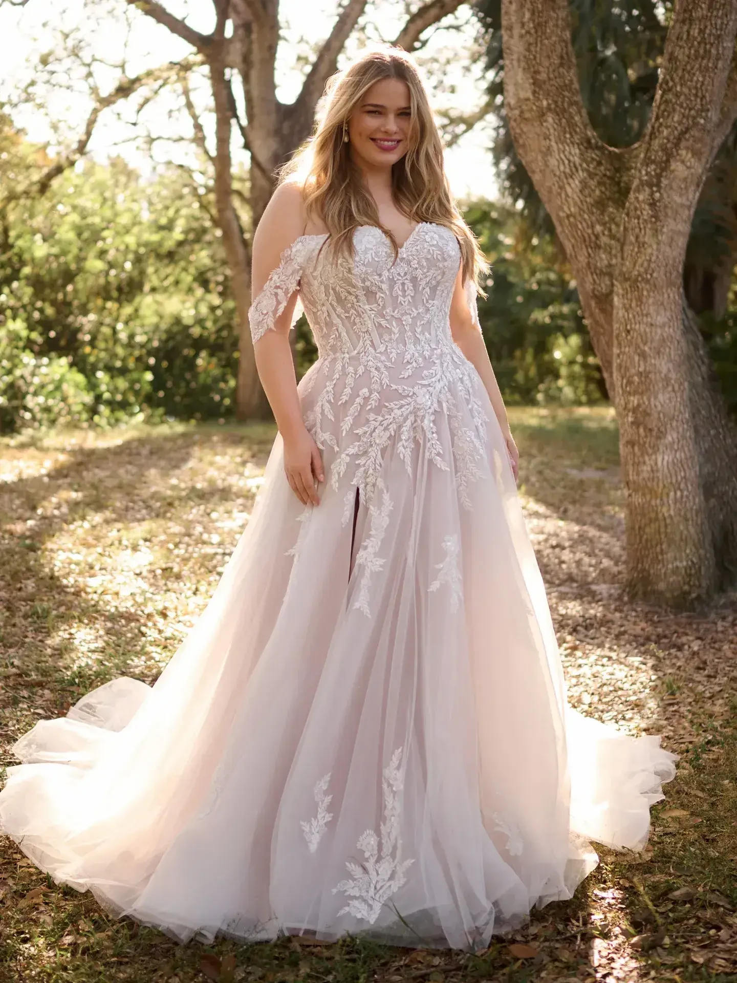 Model is wearing a sparkly A-line bridal gown with floral appliqués, a thigh-high slit, and a flowing tulle skirt